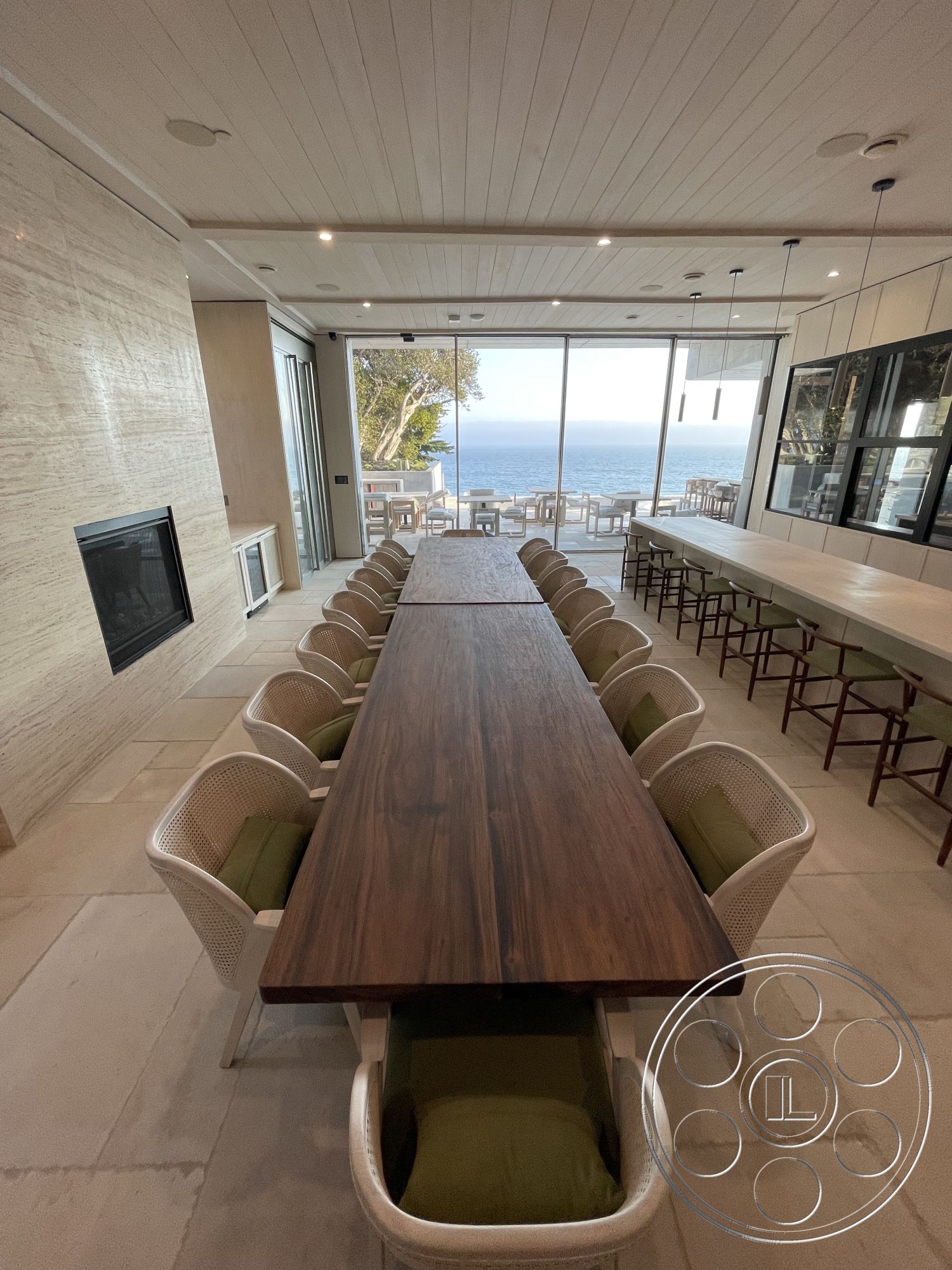 Modern 509 - seamless indoor-outdoor transition, natural wood dining table, contemporary bar stools, open-concept layout, light-colored stone flooring, interior dining area, natural light-filled space, modern pendant lighting fixtures, textured stone fireplace wall, white shiplap ceiling, upholstered dining chairs with green fabric, large glass windows with ocean view, minimalist kitchen cabinetry, luxurious coastal design elements, outdoor dining patio, spacious seating arrangement