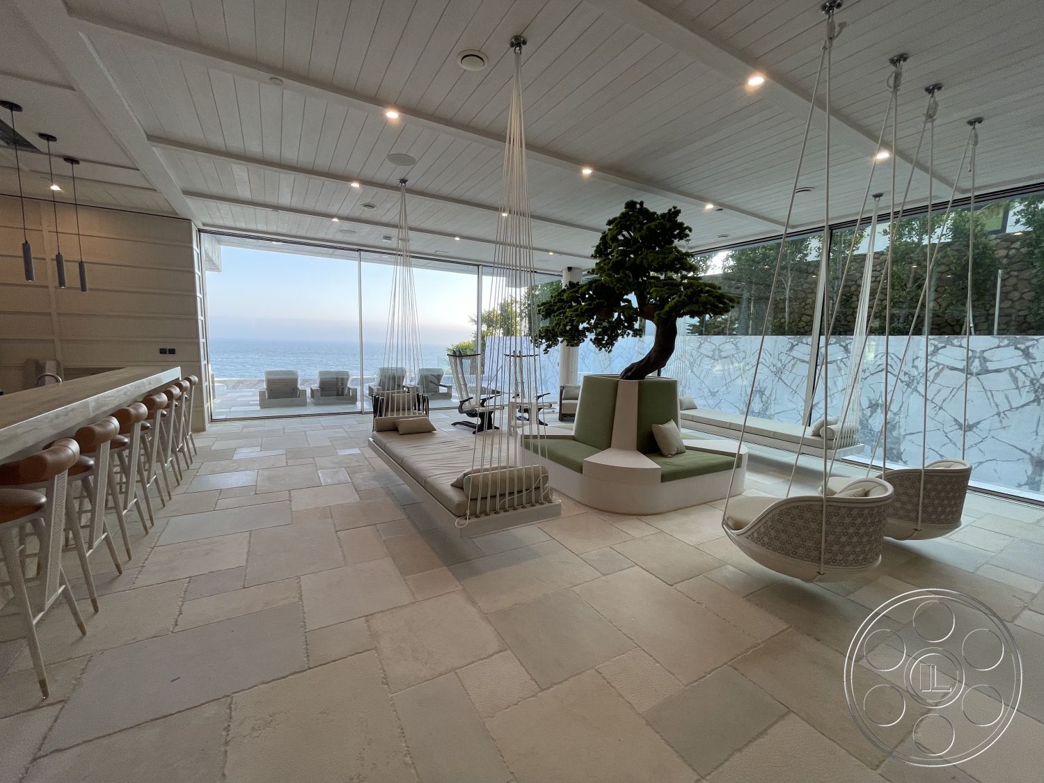 Modern 509 - ocean view, large glass windows, hanging chairs, contemporary decor, minimalist design, casual dining area, smooth white walls, stone flooring, natural light-filled space, open concept living area, textured stone accent wall, exposed wood ceiling panels, woven lounge swings, built-in green seating area, bar counter with wooden stools, indoor tree feature, swing chairs
