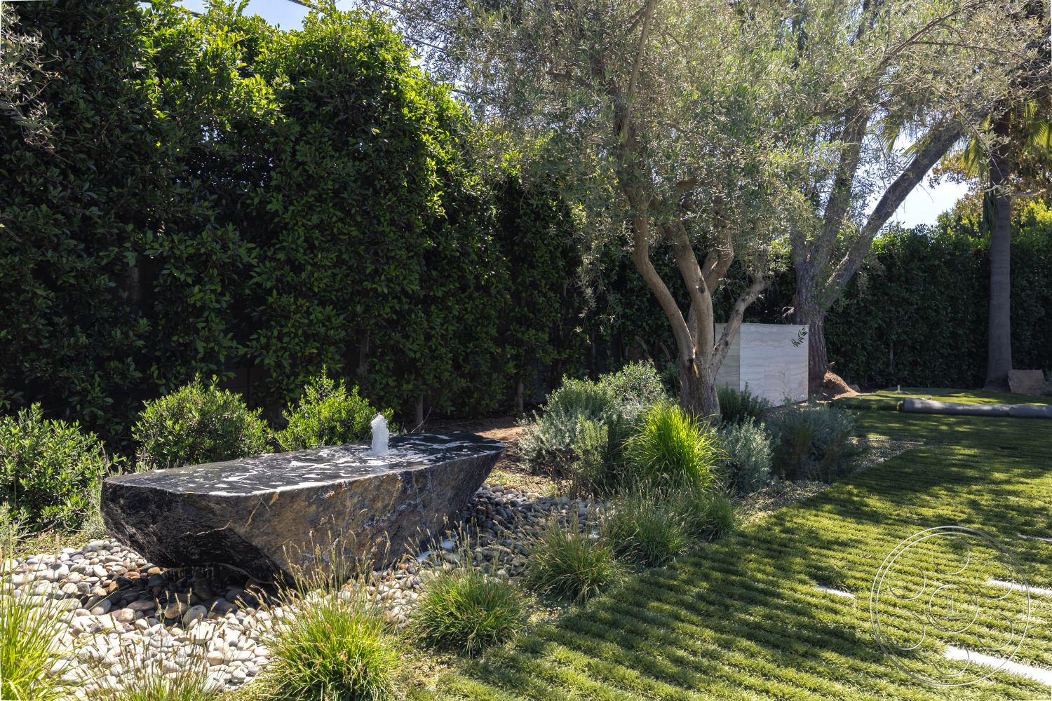 Modern 510 - fountain, low-maintenance xeriscaping, landscaped garden with native plants, exterior water fountain feature, natural stone fountain base, polished granite fountain top, gravel pathway edging, lush greenery with ornamental grasses, olive tree shading the area, concrete wall privacy screen, textured natural stone accents, stone boulder garden feature, manual irrigation system for landscaping, outdoor zen garden style, sunny backyard relaxation area, modern minimalist landscape design