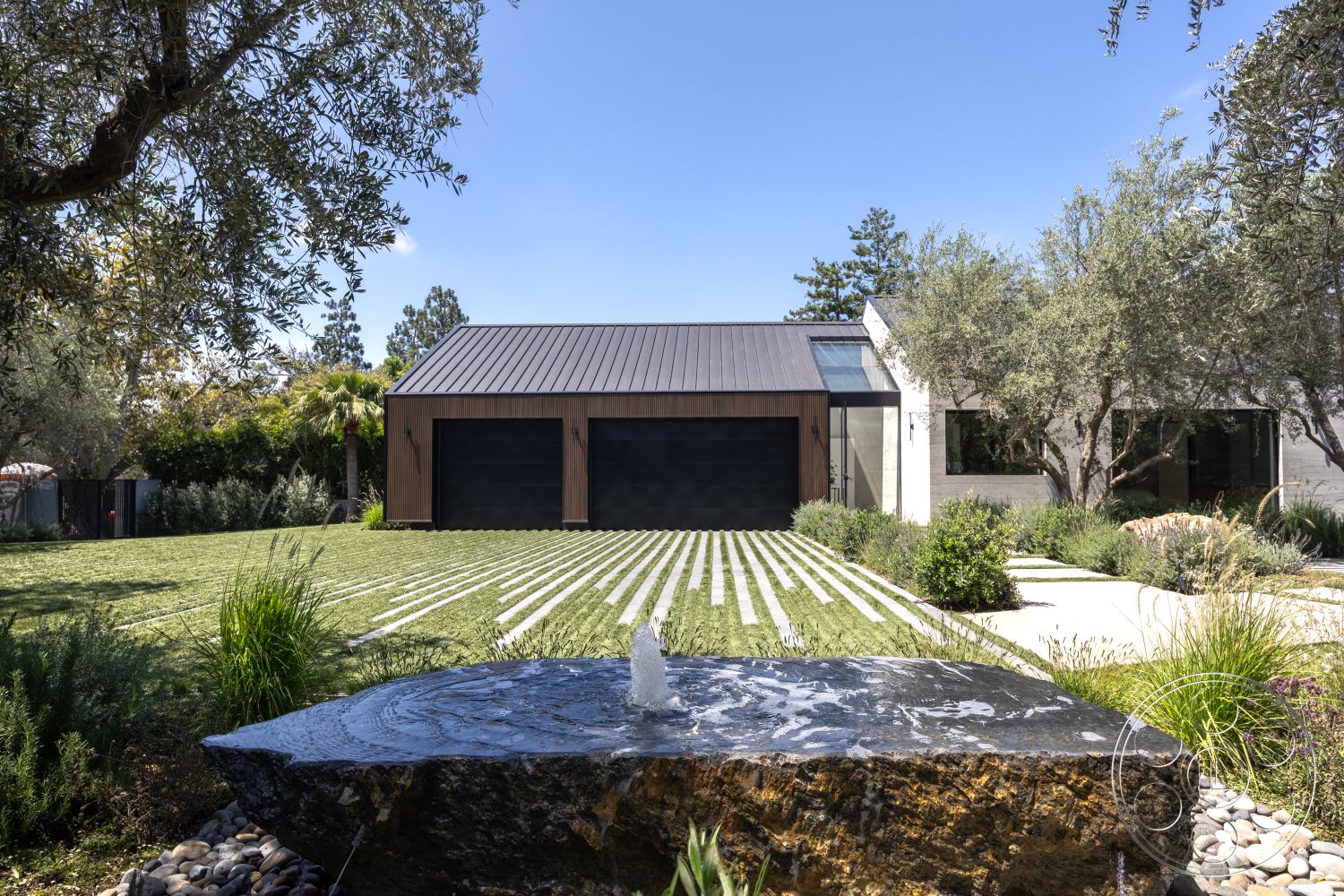 Modern 510 - exterior, surrounding greenery, modern architectural style, open outdoor space, concrete driveway, minimalist landscaping, outdoor water feature, smooth concrete walls, black metal roofing, wood panel garage doors, landscaped yard with grass, natural stone fountain, textured stone base, striped grass pattern driveway, bushes and ornamental plants, elevated design with clean lines