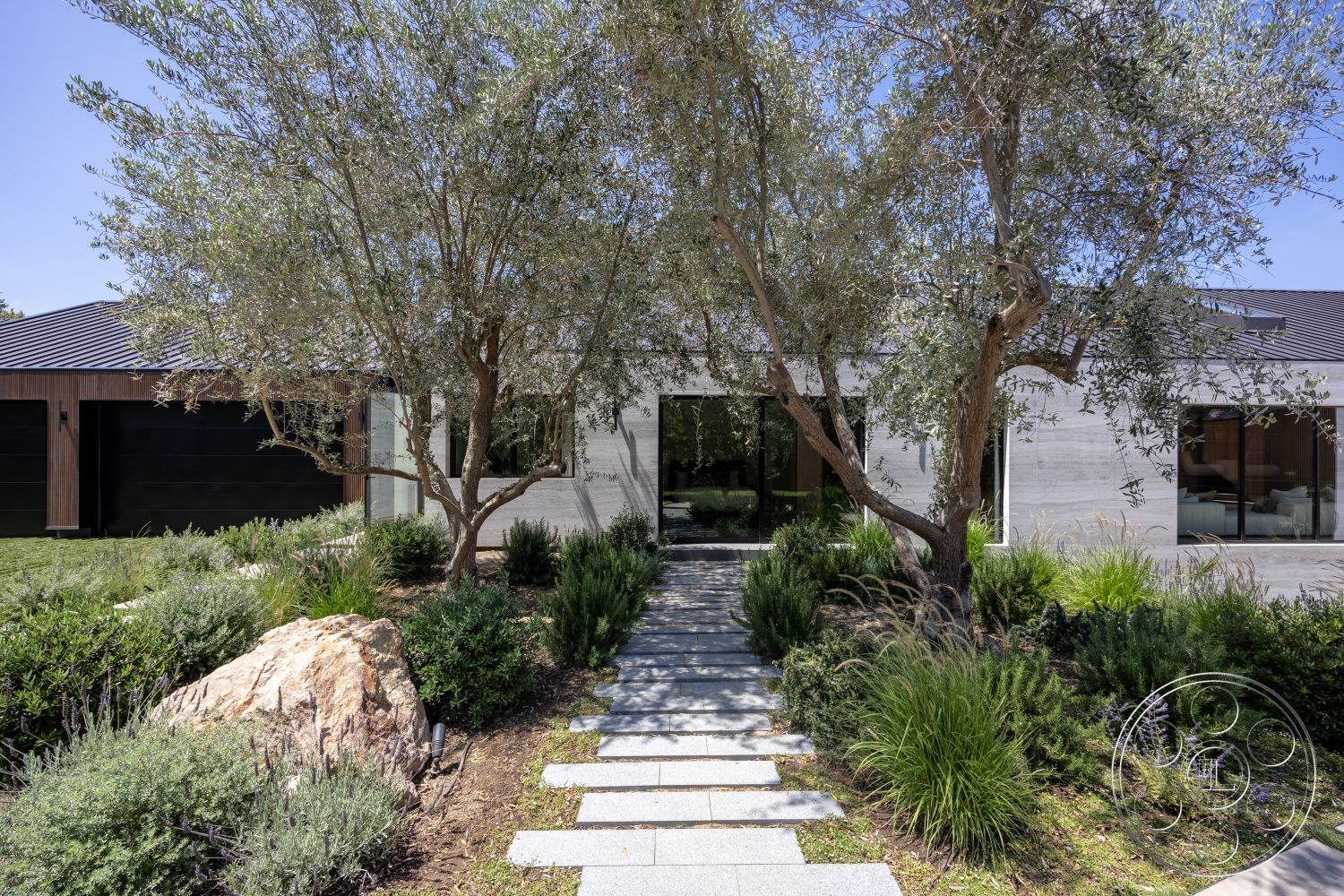 Modern 510 - exterior concrete wall, exterior concrete pathway, exterior olive trees, exterior natural stone boulders, exterior landscaped garden with low shrubs, exterior contemporary home with large glass windows, exterior horizontal wood siding, exterior metal roof, exterior sleek modern facade, exterior softscape with native plants, exterior clear view of interior through glass walls, exterior minimalist design, exterior sunlight filtering through tree leaves, exterior open space around the home, exterior seamless transition between landscape and architecture