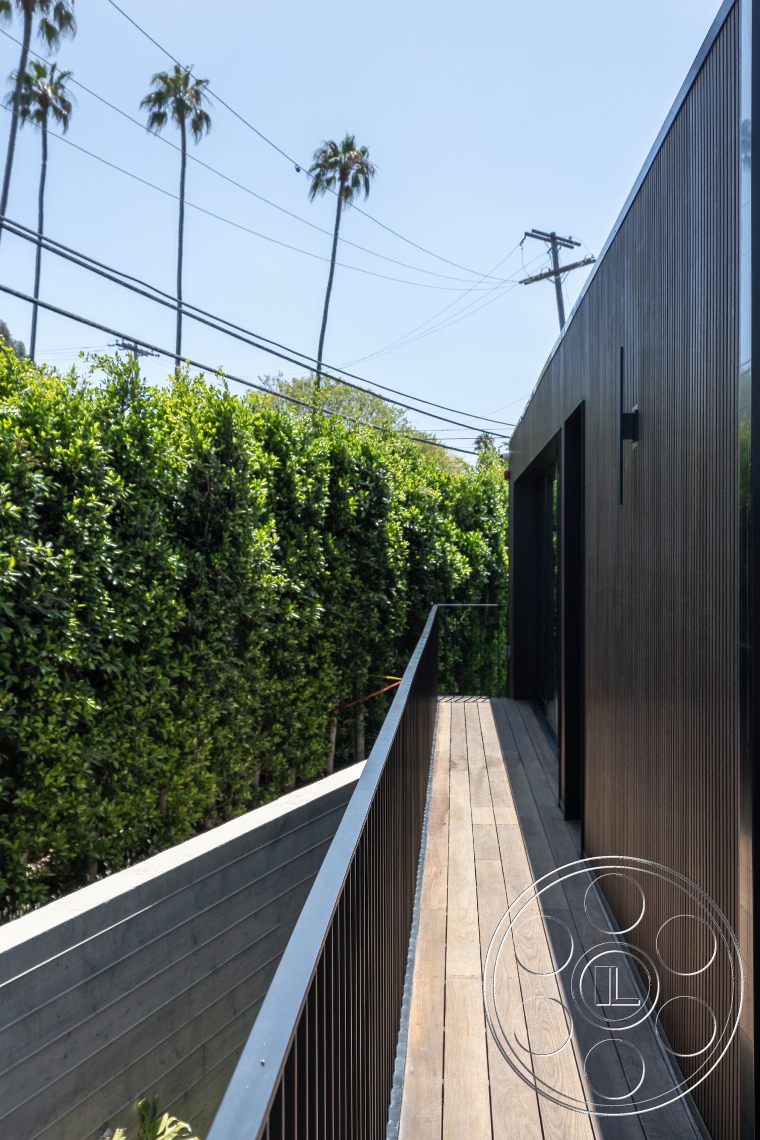 Modern 510 - balcony, modern architecture, urban setting, black metal railing, minimalist design elements, outdoor corridor, palm tree view, natural light exposure, vertical wood siding, wood deck walkway, landscaped hedge fence, interior-exterior transition space