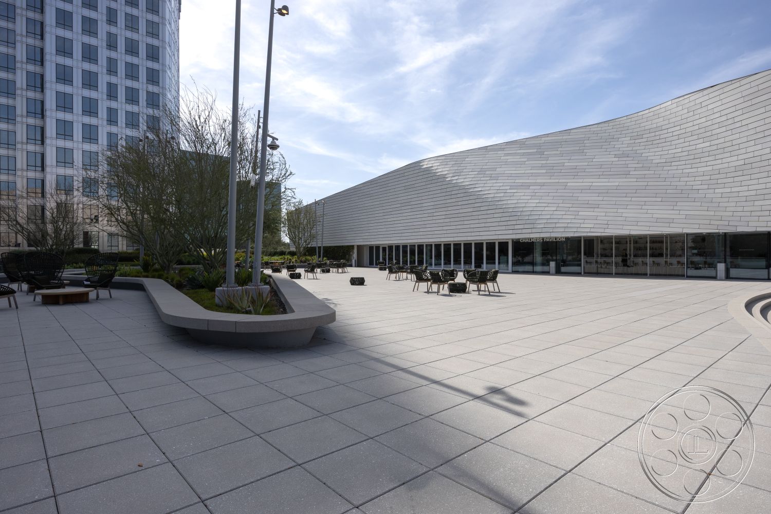 Museum 4 - urban setting with high-rise buildings, outdoor lounge area with modern chairs, exterior plaza with concrete pavers, curved concrete seating platform, landscaped green areas surrounding seating, modern architectural design with curved roof, large glass windows on pavilion, plaza with minimalist aesthetic, softscape with small trees and plants, natural light flooding into pavilion, integrated outdoor lighting fixtures, spacious outdoor gathering space, accessible design with smooth surfaces