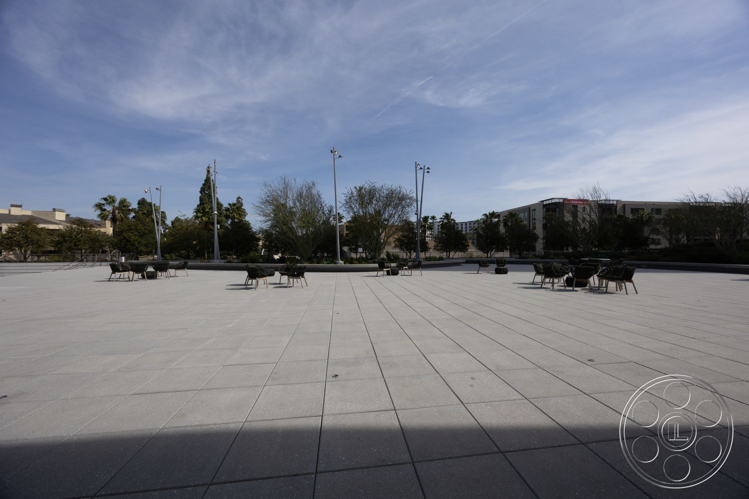 Museum 4 - urban setting, open public space, exterior concrete patio flooring, rectangular outdoor seating area, smooth finish concrete surface, dining chairs with cushions, landscape trees in background, decorative light poles, low landscape planters, large sky view