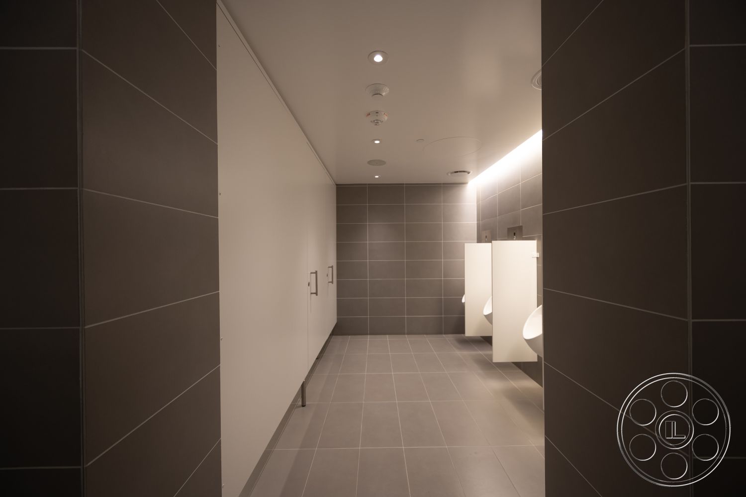 Museum 4 - textured wall surface, interior bathroom, white painted ceiling, flush mount ceiling lights, clean lines and geometric shapes, ambient LED lighting, modern restroom design, gray porcelain tile walls, gray porcelain tile flooring, minimalist wall-mounted sinks, handicap-accessible restroom features, stainless steel grab bars, modern stall partitions, functional restroom design