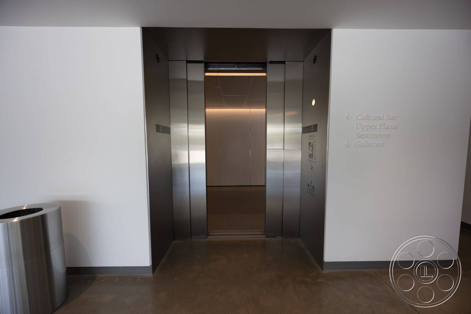 Museum 4 - polished concrete floor, soft ambient lighting, modern minimalist design, interior elevator lobby, stainless steel elevator doors, contemporary architectural details, smooth white painted walls, signage with directional arrows, trash bin with stainless steel finish