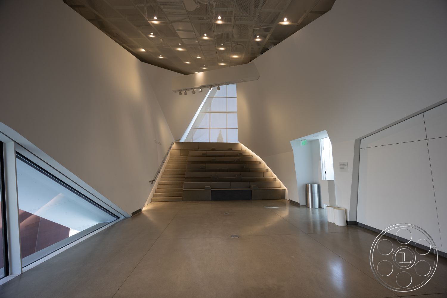 Museum 4 - neutral color palette, open floor plan, functional design, industrial lighting fixtures, large floor-to-ceiling windows, contemporary aesthetic, white plaster walls, minimalist interior, exposed concrete flooring, urban setting interior, seamless transition between spaces, creative workspace environment, natural light optimization, angular ceiling design, stepped seating area, modern architectural space, steel support elements, geometric shapes and forms, indoor amphitheater