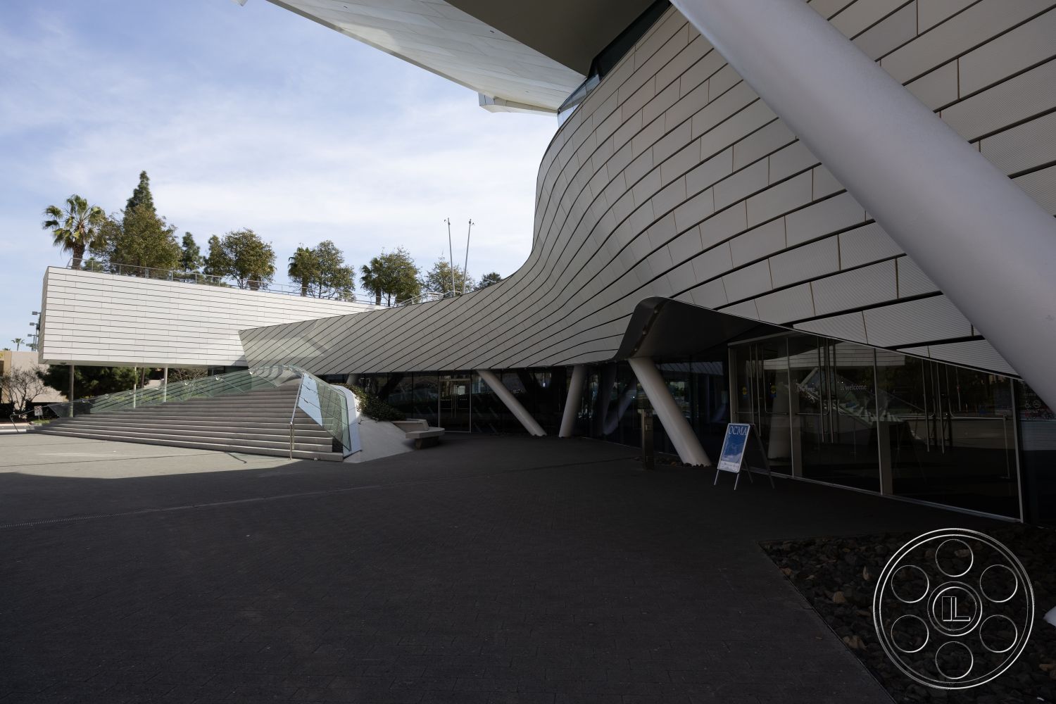 Museum 4 - exterior, surrounding greenery, urban environment, textured stone pathway, modernist design elements, curved architectural forms, glass entrance doors, metal cladding facade, landscaped outdoor seating area, geometric stair design, concrete steps with glass railing, open plaza setting, overhanging roof structure, naturally illuminated interior, visually striking architectural lines, multilevel exterior, sculptural building elements