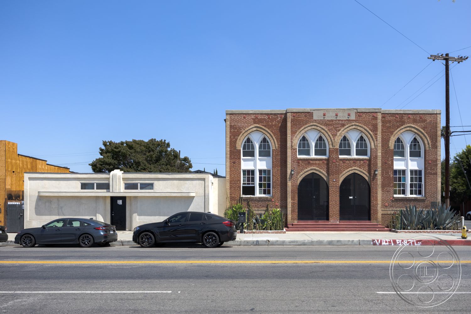 Studio 100 - concrete steps, red brick walls, flat roof design, asphalt parking area, exterior brick facade, urban street setting, arched window shapes, double wooden doors, decorative cornice details, modern concrete building element, landscaped entryway with succulents, two-story building structure, large windows with metal grilles, adjacent stucco finish building, mixed-use architectural style