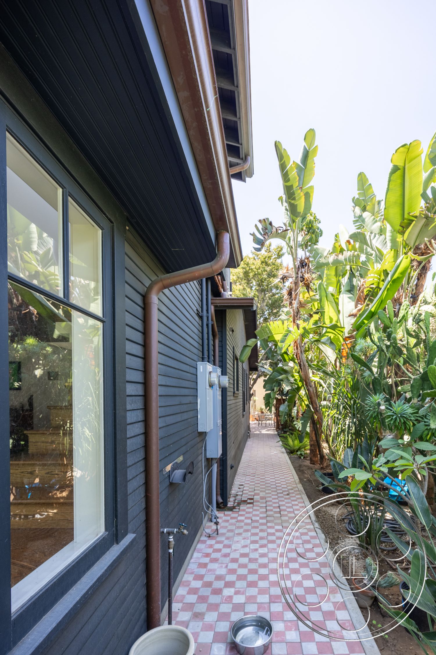 Studio 100 - textured wall surface, wood-framed windows, sunny outdoor setting, concrete foundation, landscaped garden with tropical plants, lush plant foliage, exterior black wooden siding, red and white checkered tile pathway, metal downspout, electrical meter box mounted on wall, outdoor drainage system, narrow garden walkway