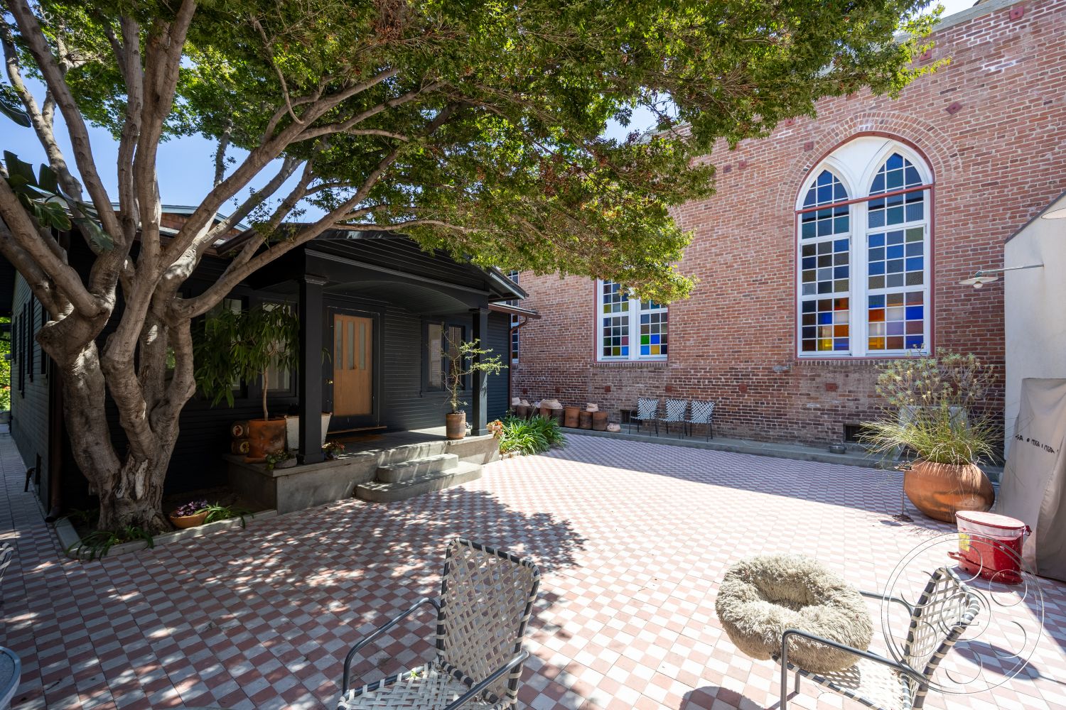 Studio 100 - exterior, exposed brick wall, concrete steps, fenced property, stained glass windows, concrete planter, wooden front door, urban residential setting, modern outdoor seating, textured concrete wall, landscaped garden area, sunlit courtyard, urban courtyard, checkerboard tile patio flooring, large shade tree, decorative outdoor pots, black wooden exterior, adjacent historical building