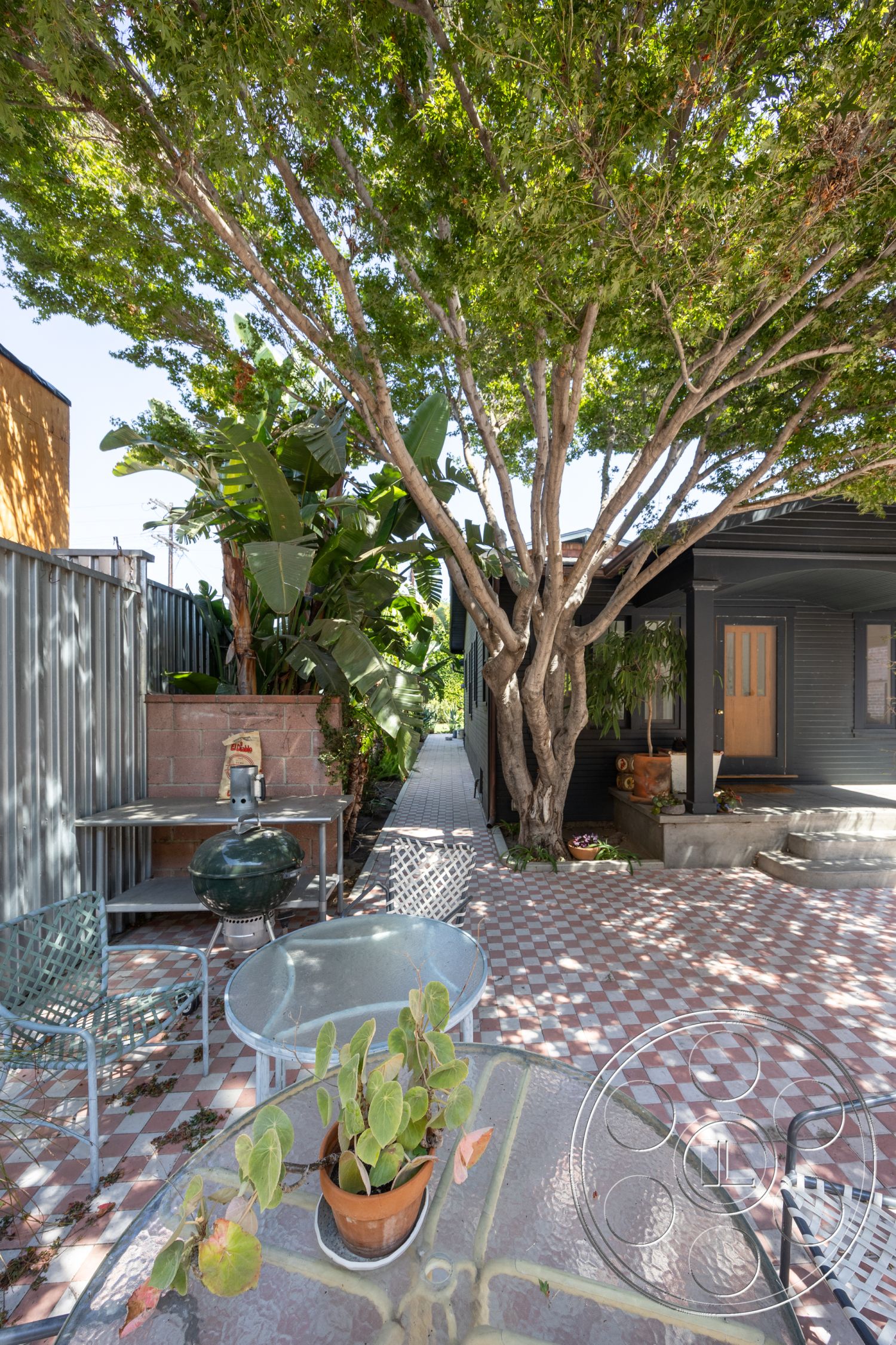 Studio 100 - decorative potted plants, urban backyard, landscaped garden setting, brick garden wall, exterior patio with patterned tile flooring, red and white checkered patio tiles, metal outdoor dining chairs, glass-top outdoor dining table, mature shade trees, pathway leading to front door, modern home with dark exterior siding, grill area with outdoor cooking space, lush tropical foliage surrounding patio