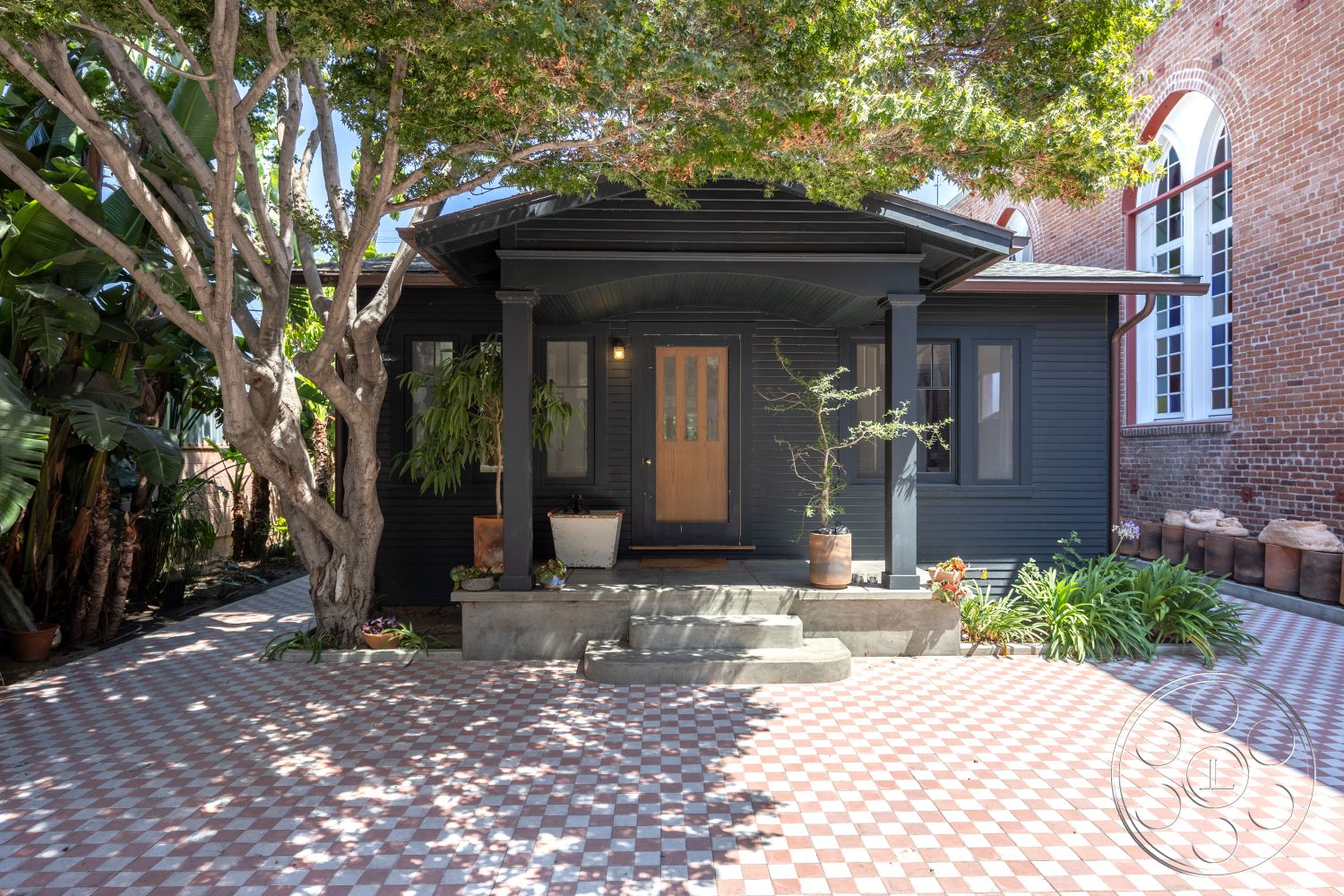 Studio 100 - exterior, lush tropical landscaping, dark wood paneling, colonial architectural style, chequered tile patio flooring, concrete porch steps, brick building wall, exposed tree canopy, traditional front porch, wood front door with glass insert, potted plants at entrance, multicolored tile patterns, stepped concrete foundation, scenic garden setting, outdoor decorative planters