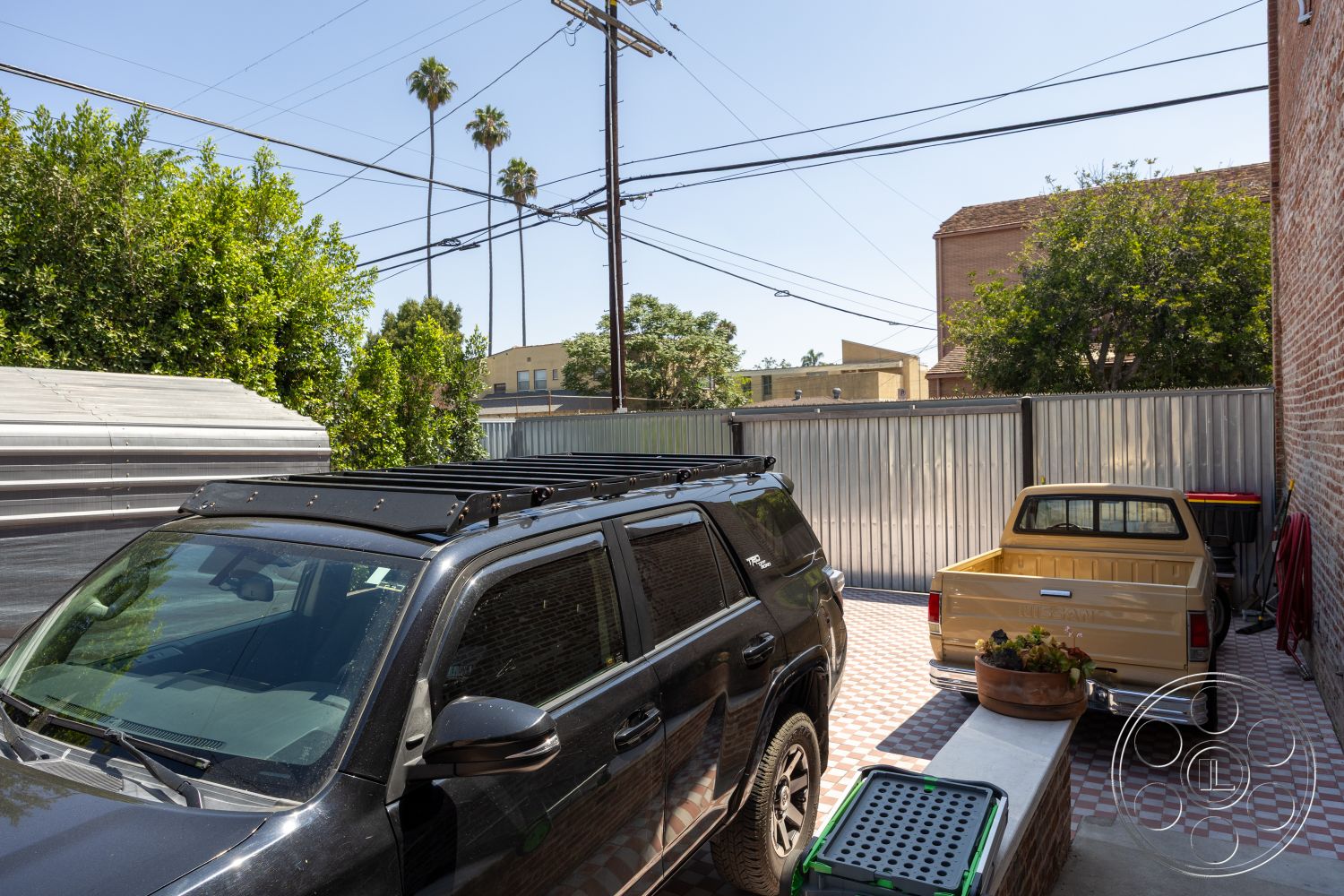 Studio 100 - exterior, exposed brick wall, brick wall, metal fence, residential backyard, patterned tile flooring, surrounding cityscape, urban parking area, concrete outdoor seating, checkerboard patio tiles, landscaping with palm trees, industrial-style outdoor space, vehicle parking area, garden planter, metal storage shed, bright sunny setting