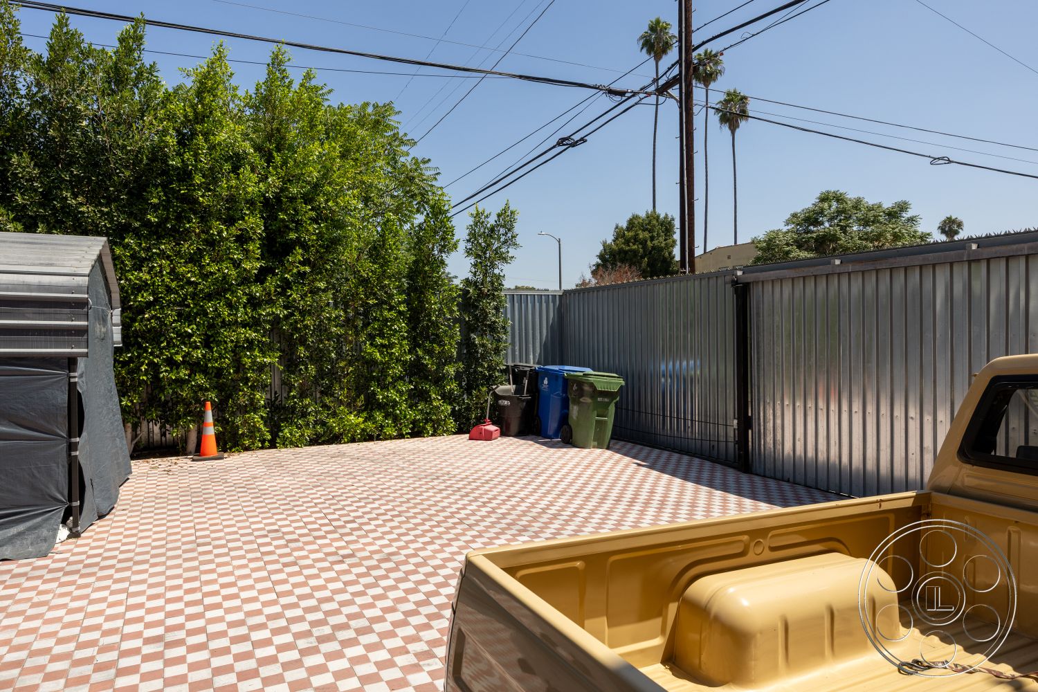 Studio 100 - outdoor space, landscaped yard, utility area, metal fencing, tropical foliage, lush greenery backdrop, sunny environment, brick paver driveway, functional exterior space, checkerboard tile flooring, exterior parking area, concrete wall dividers, city setting, industrial-style container storage, urban outdoor parking space, colorful trash bins, tarmac outdoor flooring, outdoor utility shed