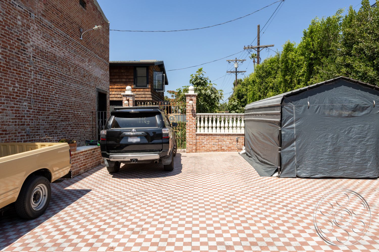 Studio 100 - exposed brick walls, brick garden wall, open-air parking area, urban outdoor setting, checkered tile driveway flooring, metal fence gates, wooden shed structure, contemporary home exterior, concrete patio slab, suburban residential environment, two-story home adjacent, landscaped backyard view, sunlit driveway area