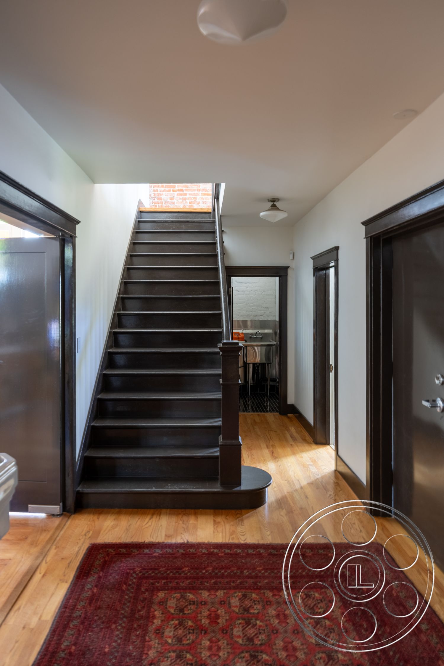 Studio 100 - neutral color palette, decorative area rug, vaulted ceiling, natural light from skylight, white painted walls, exposed brick accent wall, hardwood foyer flooring, black wooden staircase, dark wood paneling on doors, traditional wooden handrail, interior stairwell, modern ceiling light fixture, open-concept hallway, industrial door hardware, pedestrian-friendly ground floor layout