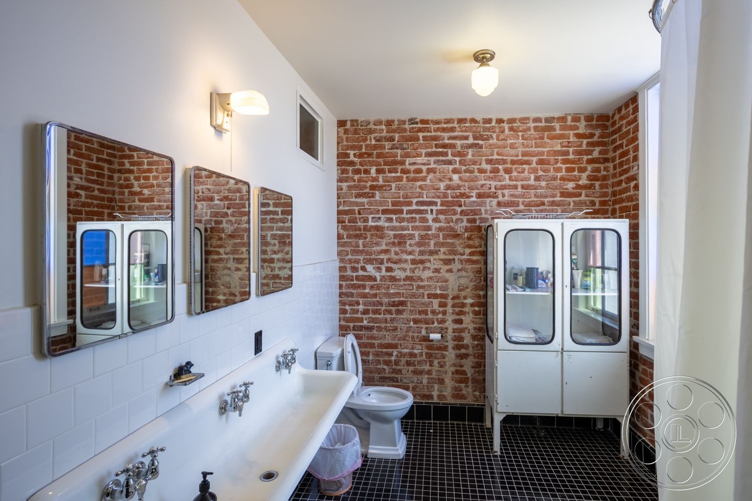 Studio 100 - minimalist bathroom design, natural light from window, interior bathroom space, exposed brick accent wall, white subway tile shower walls, black checkered tile flooring, double vanity with porcelain sink, framed mirrors in bathroom, vintage-style ceiling light fixture, metal cabinet for bathroom storage