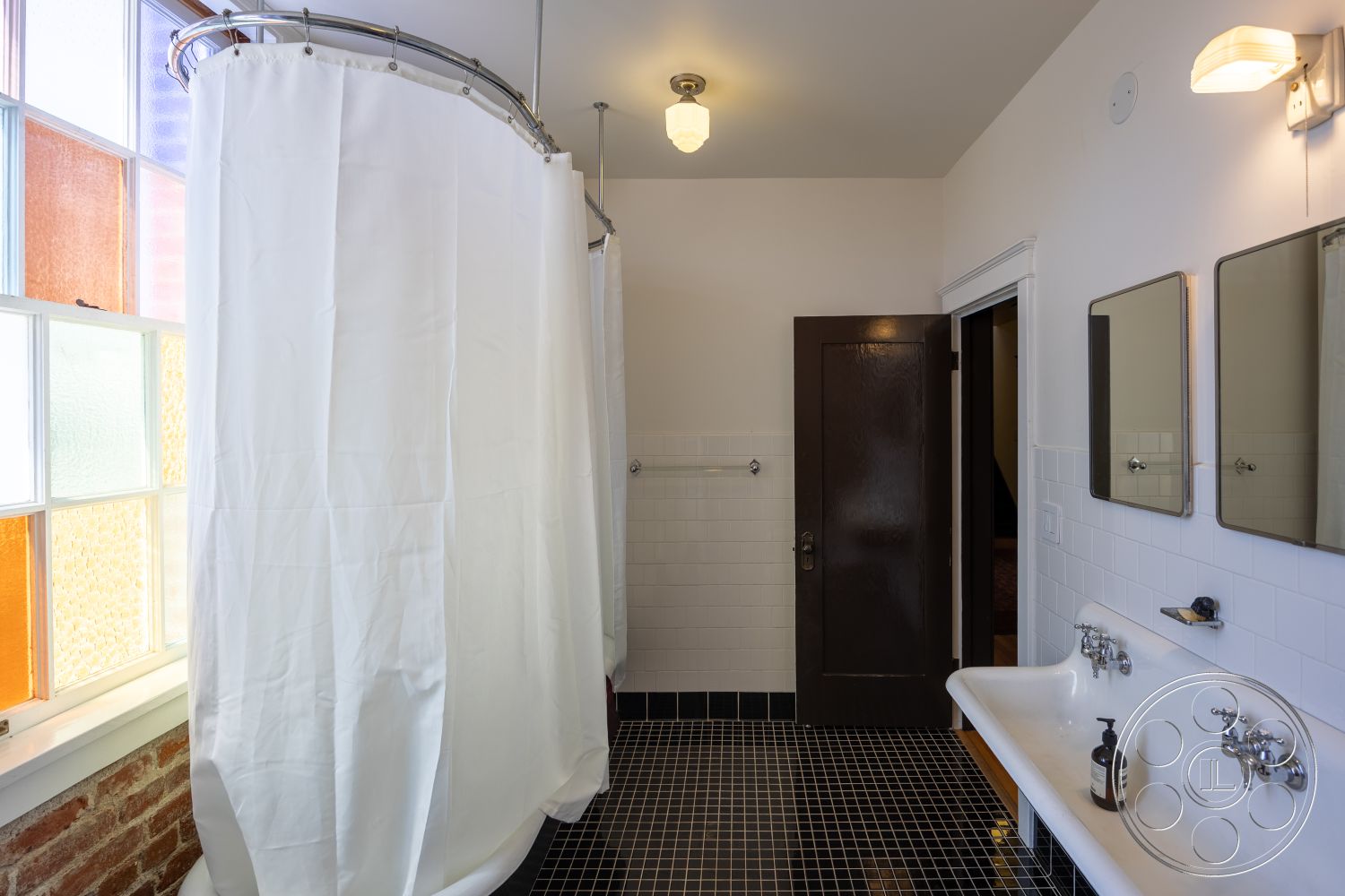 Studio 100 - black tile flooring, dark wood door frame, exposed brick wall interior, white tiled bathroom walls, curved shower curtain rod, double sink basin with classic faucets, retro-style wall-mounted mirrors, colorful leaded glass windows, industrial-style lighting fixture, minimalist bathroom space, separate shower area with curtain, functionally designed wash area, pre-war bathroom design