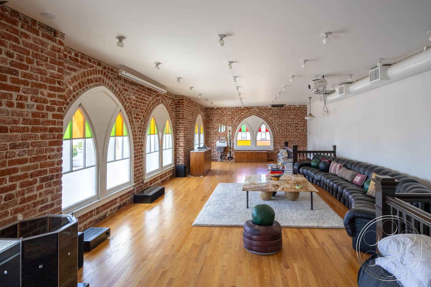 Studio 100 - cozy seating arrangement, neutral area rug, textured plaster ceiling, hardwood living room floors, minimalist decor elements, open floor plan layout, exposed brick accent wall, urban loft setting, industrial ceiling lights, contemporary home office area, arched windows with stained glass, modern leather sectional sofa, wooden coffee table with natural finish, high ceilings with visible ductwork, colorful window treatments