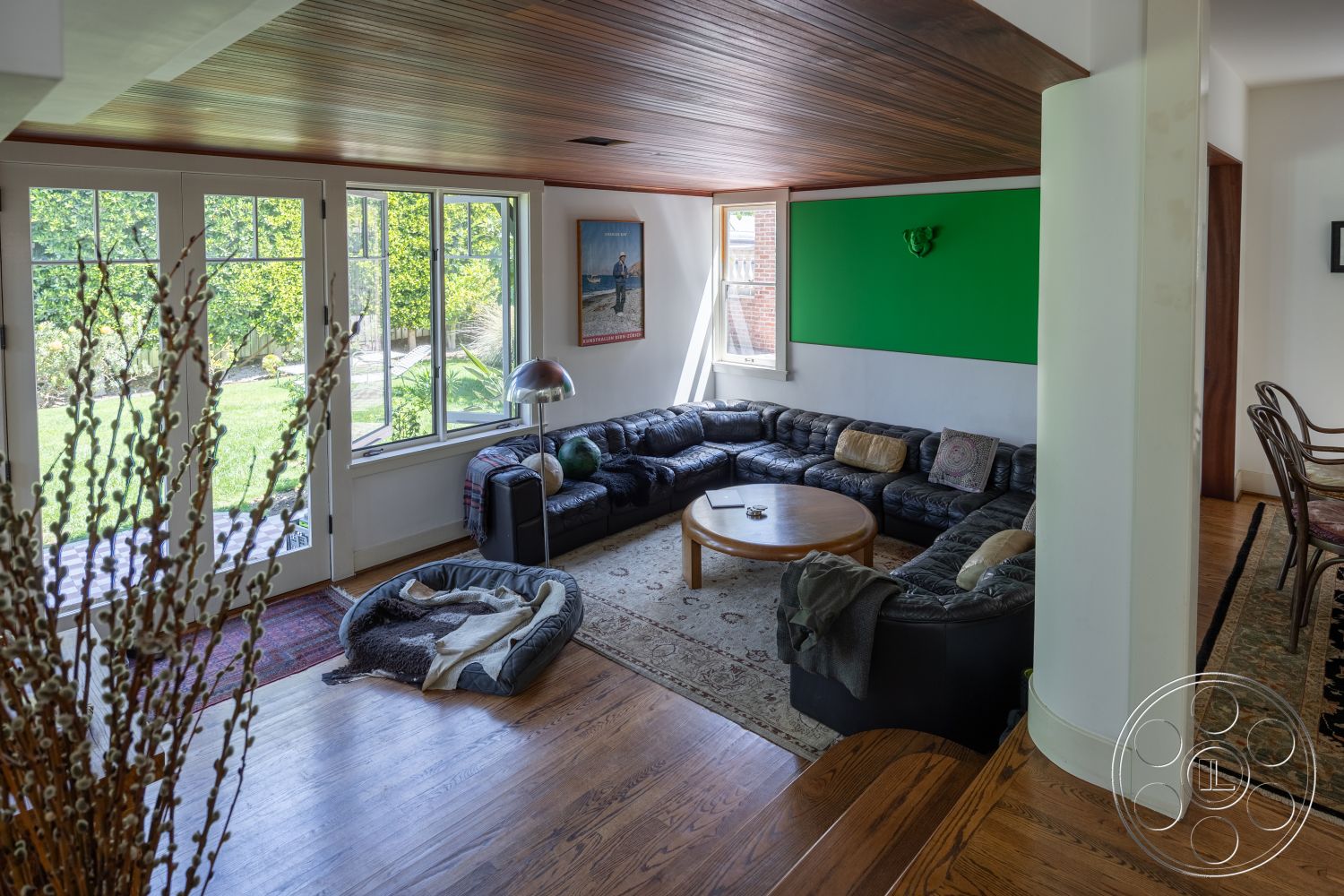 Studio 100 - textured area rug, open living space, green accent wall, round wooden coffee table, modern floor lamp, hardwood living room floors, wood beam ceiling, large windows with garden view, exposed brick accent wall, black leather sectional sofa