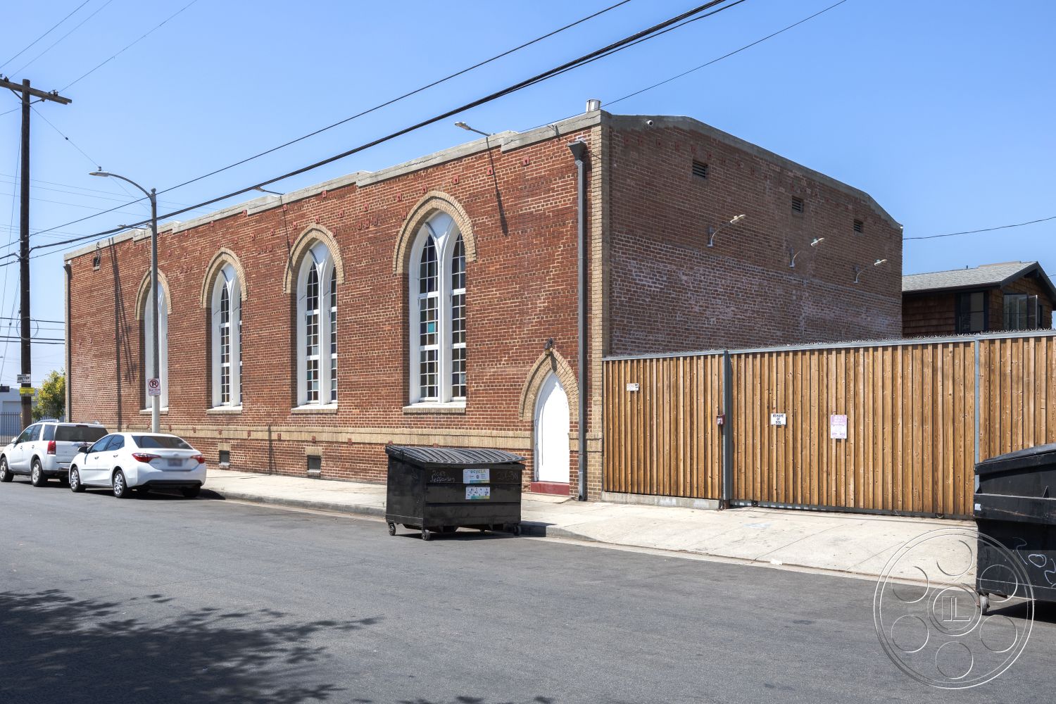 Studio 100 - urban setting, large multi-pane windows, vehicular access, concrete sidewalk, flat roof construction, exposed brick exterior walls, arched window frames, gabled roof profile, wooden fence enclosure, historic architectural style, industrial neighborhood, cylindrical chimney stack, contrast between brick and wood materials, adjacent urban street view