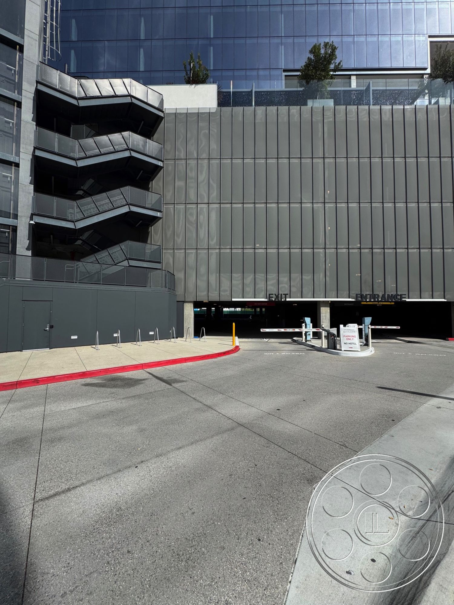 Office 121 - urban setting, modern architectural style, curved driveway, textured concrete wall, flat roof design, concrete driveway surface, floor-to-ceiling glass windows, commercial building exterior, exposed steel framework, concrete parking structure, metallic exterior siding, glass facade wall, zigzag fire escape stairs, red curb detailing, multi-level parking entrance, interior parking area