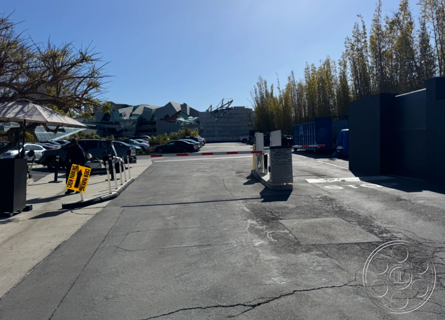 Hayden Tract - interior-exterior flow, exposed structural elements, surrounding urban environment, urban landscape setting, metal security gate, asphalt parking lot surface, contemporary architectural forms, commercial parking area, landscaped tree border, bay window architectural detail, flat-roofed building silhouette, horizontal wood slat fencing, open air parking structure, utility shed structure, faded pavement markings, blue delivery truck parking, notices on yellow signage, barrier arm gate