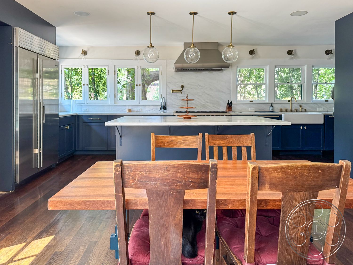 Craftsman 20 - open floor plan, stainless steel refrigerator, lush greenery visible through windows, modern kitchen appliances, functional kitchen layout, interior kitchen with marble countertops, minimalist kitchen design, hardwood flooring in kitchen area, white subway tile backsplash, dark blue kitchen cabinets, glass pendant lights over kitchen island, double sink with brass faucet, decorative wood dining table, multiple windows for natural light, cozy dining space with upholstered chairs, bright, airy atmosphere