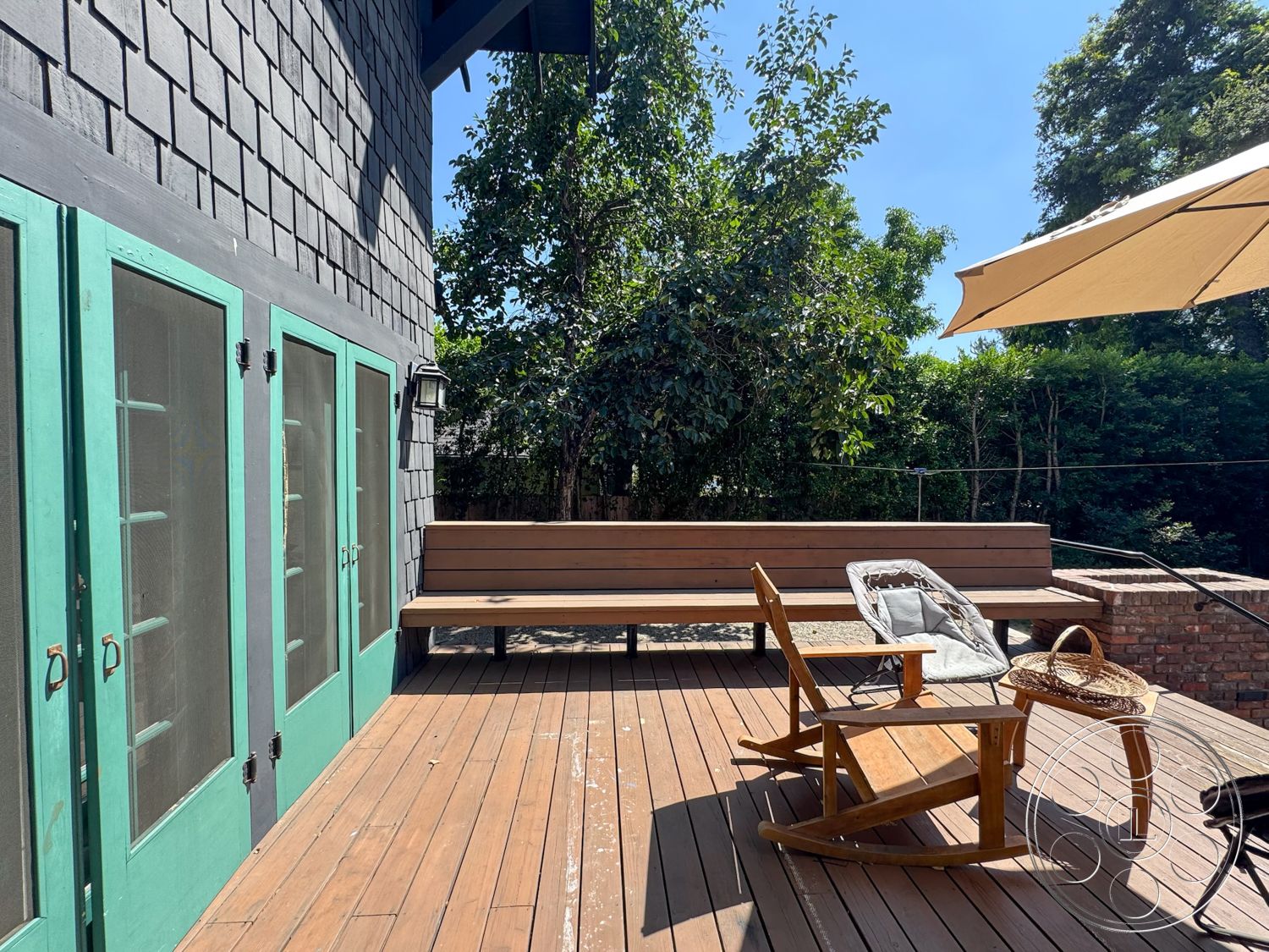 Craftsman 20 - wooden beams, modern rustic style, wooden deck flooring, lush garden setting, exterior patio deck, gray shingle exterior wall, turquoise painted double doors, built-in wooden bench seating, outdoor rocking chair, brick low wall border, green outdoor umbrella, sunlit backyard, casual outdoor lounge area, direct access to garden