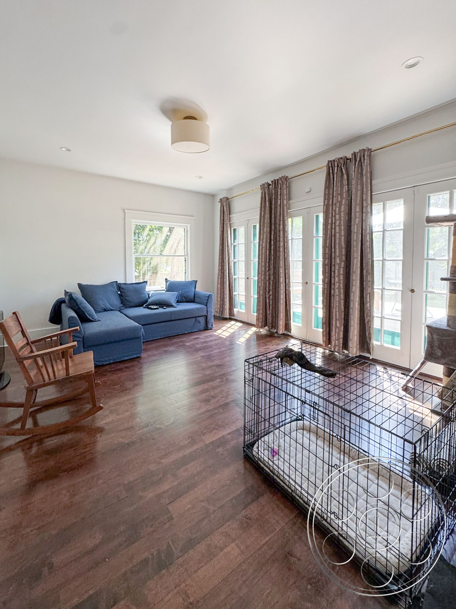 Craftsman 20 - French doors, modern light fixture, textured fabric curtains, curtained windows, wooden rocking chair, hardwood living room floors, natural light interior, open floor plan layout, white painted walls, blue upholstery sofa, metal pet crate, coastal decor accents, homey living space