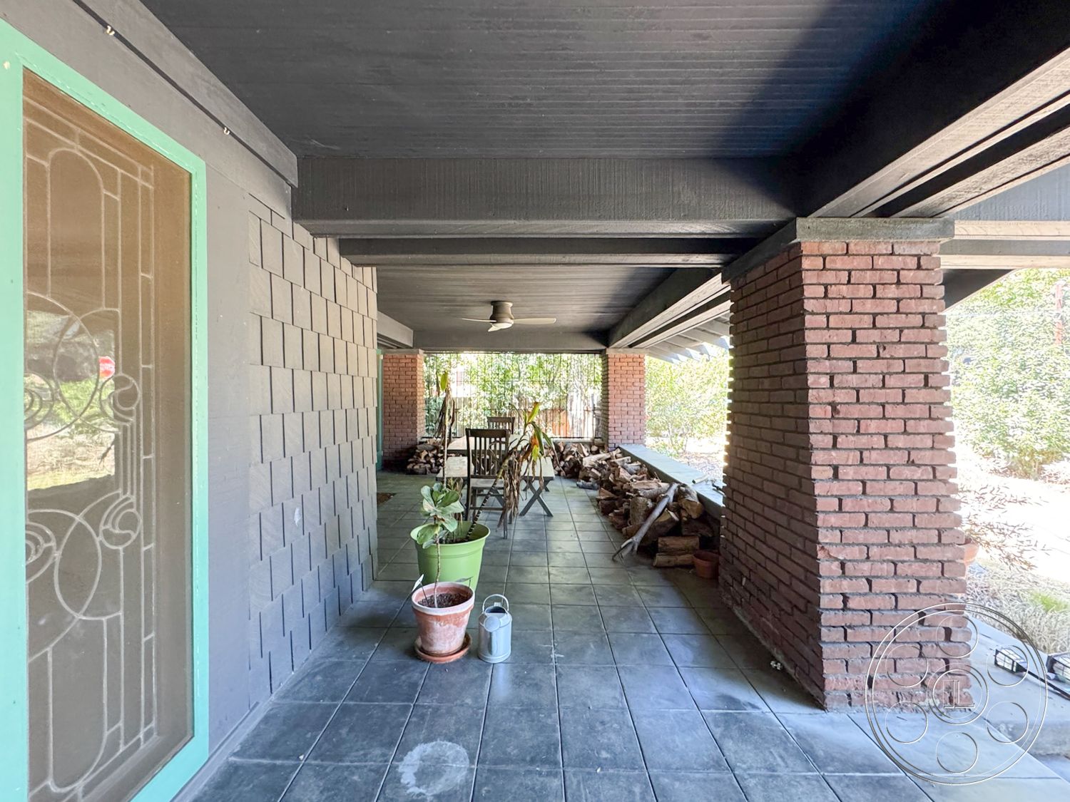 Craftsman 20 - natural landscaping, covered porch, garden setting, glass panel door, outdoor living area, planter pots, slate tile flooring, roof overhang, exposed brick accent wall, wooden seating area, exterior entryway, brick column supports, shingle siding wall treatment, black painted wood ceiling, textured wood beam detailing