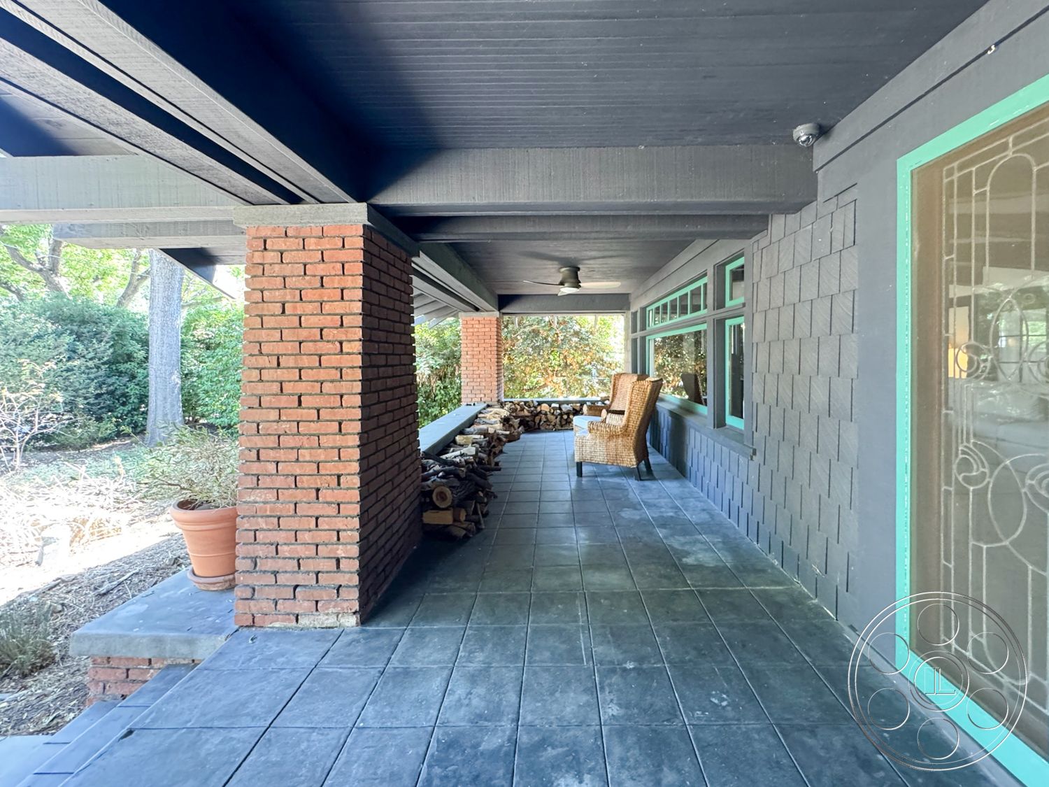 Craftsman 20 - exterior landscaped garden setting, exterior wood beam ceiling, exterior concrete patio flooring, exterior brick support columns, exterior gray shingle wall treatment, exterior large windows with green trim, exterior covered porch area, exterior stacked stone wood storage, exterior potted plant decoration, exterior fans for outdoor cooling, exterior natural foliage surroundings, exterior rustic outdoor seating area