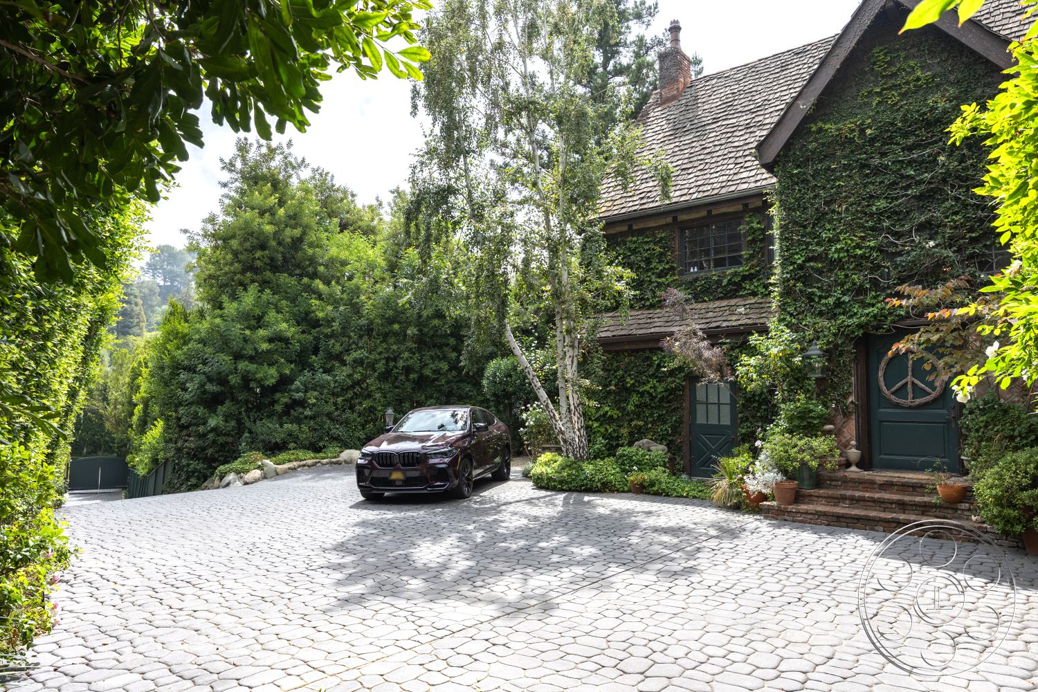 Mansion 2 - exterior, arched doorways, natural stone path, ornamental shrubs, cobblestone driveway, two-story home, urban garden setting, landscaped garden patio, exposed brick exterior walls, dark wood shingles roof, green ivy-covered facade, wooden double front doors, stone planter walls, hardwood trim elements, detailed wood window casings, rich green surrounding foliage, rustic cottage style architecture, decorative wooden accents