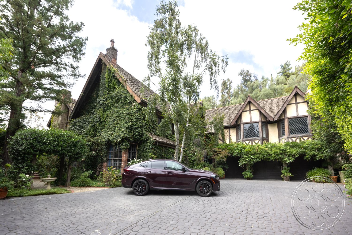 Mansion 2 - cobblestone driveway, exposed brick chimney, natural stone walkway, rustic outdoor seating area, landscaped garden with hedges, exterior stone wall with ivy, timber-framed Tudor-style house, sloped asphalt roof, arched windows with diamond lattice, paved courtyard entrance, dark wood door with window details, surrounding pine trees, lush greenery in backyard, decorative exterior gables, modern car parked on driveway, front porch with stone steps, enclosed garden space, traditional wood trim detailing
