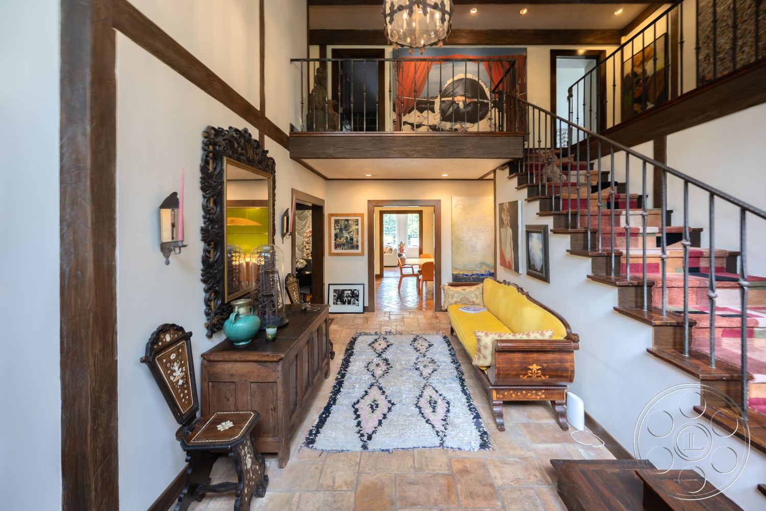 Mansion 2 - textured plaster walls, arched entryway, moody ambient lighting, colorful area rug, exposed wood ceiling beams, modern pendant light fixture, interior entrance hall, travertine tile flooring, iron stair railings, vintage wooden bench, large decorative mirror, yellow upholstered sofa, artwork gallery wall, traditional wooden side table, open-concept space design