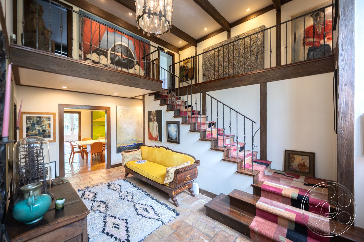 Mansion 2 - neutral color palette, stone fireplace, cozy seating arrangement, inviting entrance, textured plaster walls, modern art decor, exposed wood ceiling beams, vaulted living room ceiling, open floor plan living area, chandelier lighting fixture, rustic interior design, yellow upholstered sofa, floating stairs with carpet runner, tile entryway flooring, wooden framed doorways, large art pieces on walls, wood accent stair railing, light-filled space with large windows, multi-level foyer, natural light from entryway, urban contemporary interior