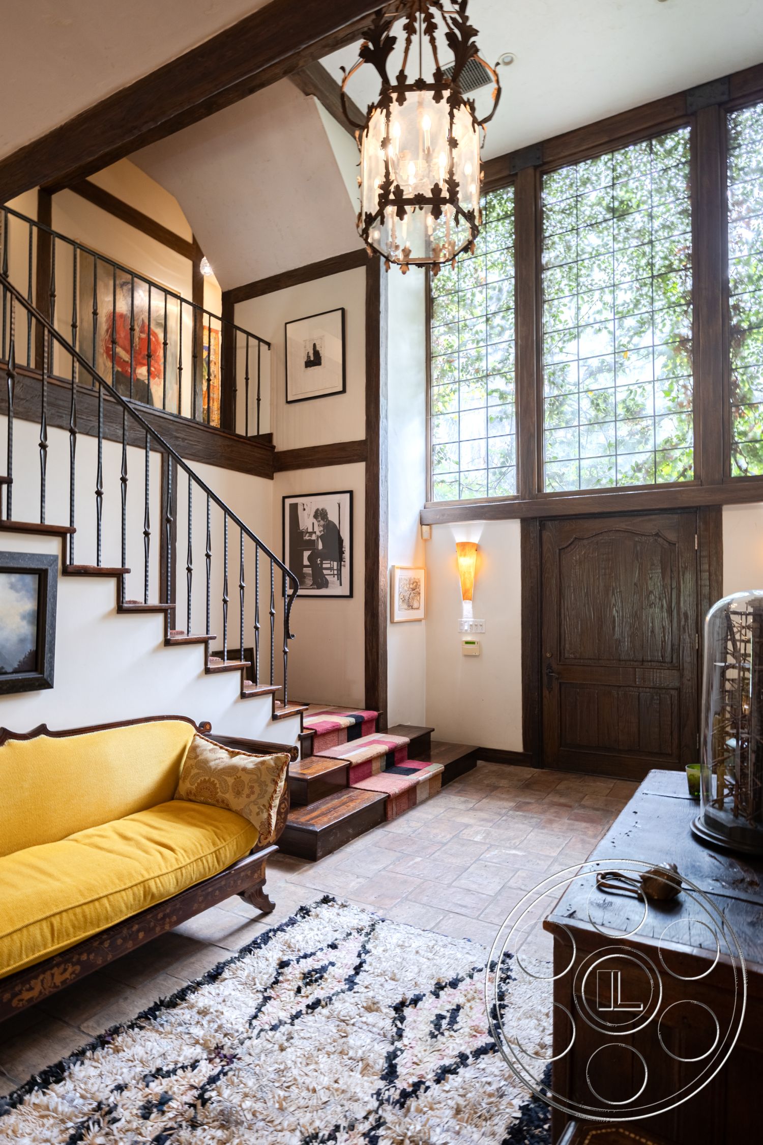 Mansion 2 - tile flooring, warm color palette, arched doorways, textured plaster walls, dark wood paneling, chandelier light fixture, home entryway, area rug with geometric pattern, wood beam ceiling, interior entrance hall, large stained glass windows, exposed wood staircase, vintage wooden front door, yellow upholstered settee, black wrought iron stair railings, mounted wall art, art deco wall sconce, richly textured wall treatments