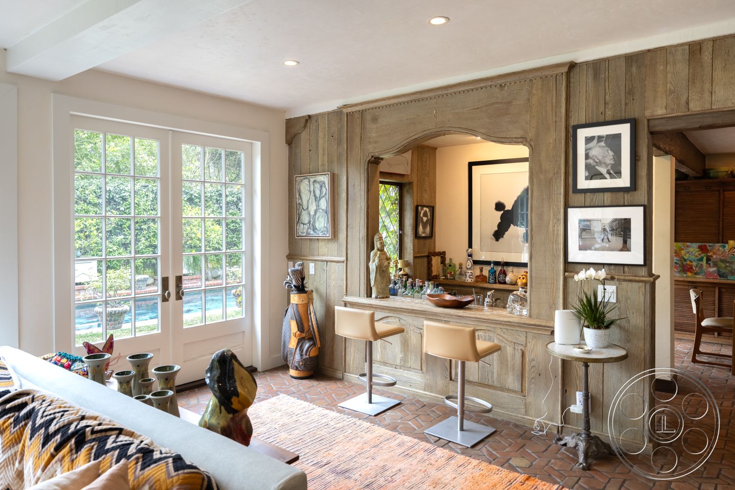Mansion 2 - neutral color palette, elegant lighting fixtures, open-concept living area, floor-to-ceiling glass doors, contemporary art pieces, exposed wood paneling interior, soft rug flooring, brick floor tiles living room, arched doorway with wood trim, rustic wooden bar counter, contemporary stool seating, art display wall with framed prints, textured fabric sofa cushions, natural light from patio doors, lush garden view from interior, artistic decor accents, handcrafted pottery decor