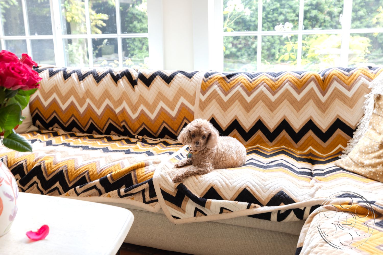 Mansion 2 - wood flooring, cozy seating area, modern coffee table, natural light from large windows, green garden view, colorful floral arrangement, interior living room, textured fabric sofa, zigzag patterned throw blanket, plush decorative pillows, small poodle dog, contemporary home decor