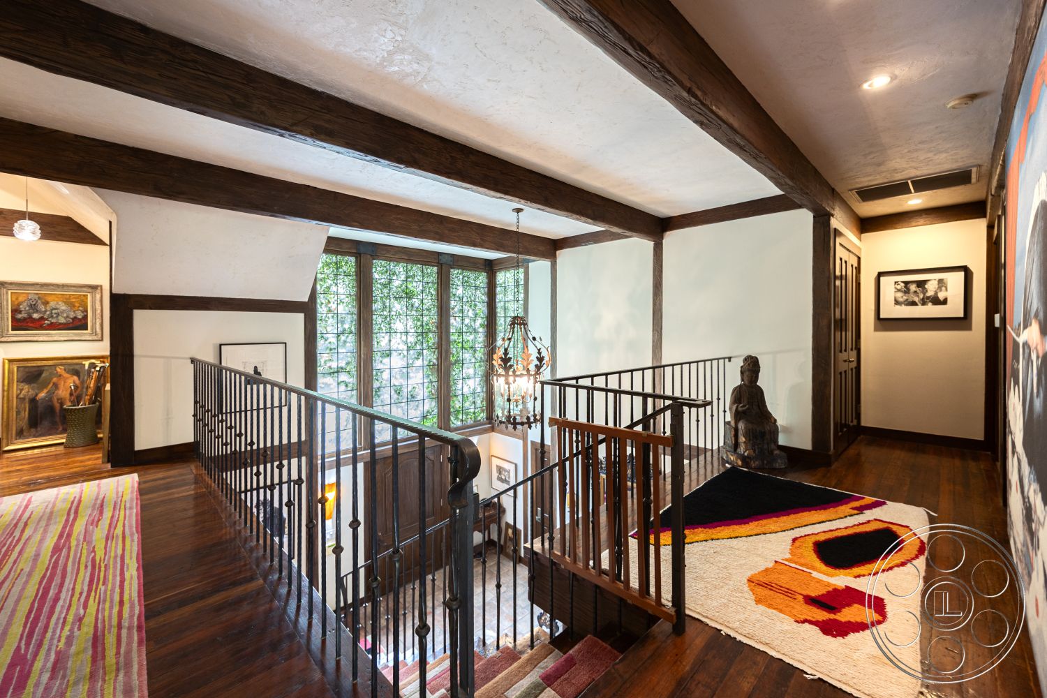 Mansion 2 - warm wood tones, textured plaster walls, exposed wood ceiling beams, natural light interior, minimalist decor accents, luxury home interior, vaulted ceiling details, artwork gallery wall, hardwood staircase flooring, iron railing staircase, large bay windows with greenery views, modern chandelier light fixture, colorful area rug with geometric patterns, contemporary home office, open landing area, elegant stair landing, sophisticated interior design
