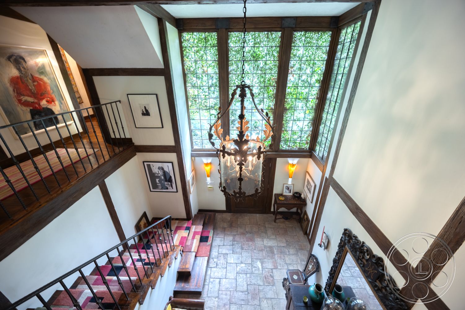 Mansion 2 - exposed wood ceiling beams interior, textured plaster walls interior, slate flooring entryway, iron staircase railing interior, arched doorway with wooden frame, square chandelier with intricate design, stone feature wall near entrance, framed artwork on walls interior, stained glass window detail, checkerboard patterned rug in foyer, dark wood paneling staircase interior, charming vintage side table near entry, decorative wall sconce lighting, open staircase leading to upper floor