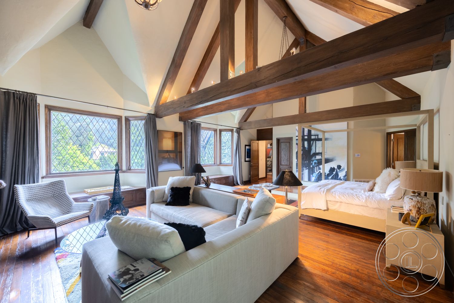 Mansion 2 - neutral color palette, cozy seating area, soft bedding, natural light from large windows, textured plaster walls, stylish coffee table, rustic furniture, modern armchair, modern art decor, contemporary lamp, mountain view setting, exposed wood ceiling beams, hardwood bedroom floors, built-in storage cabinets, open layout design, master bedroom area, wood trim details, vaulted ceiling interior, diamond-patterned window panes, wooden beams in ceiling, area rug on hardwood floors, curtains for window treatments, elegant bedside table