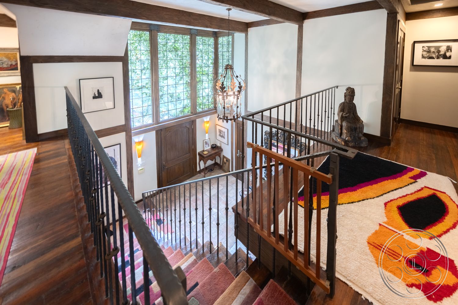 Mansion 2 - natural light from large windows, textured plaster walls, exposed wood ceiling beams, metal stair railings, wood paneling accents, hardwood staircase treads, glass block window, decorative chandelier lighting, area rug with abstract design, traditional art wall decor, vaulted ceiling in entrance hall, wood flooring in hallway, interior staircase landing, open concept landing area, home entryway with art display
