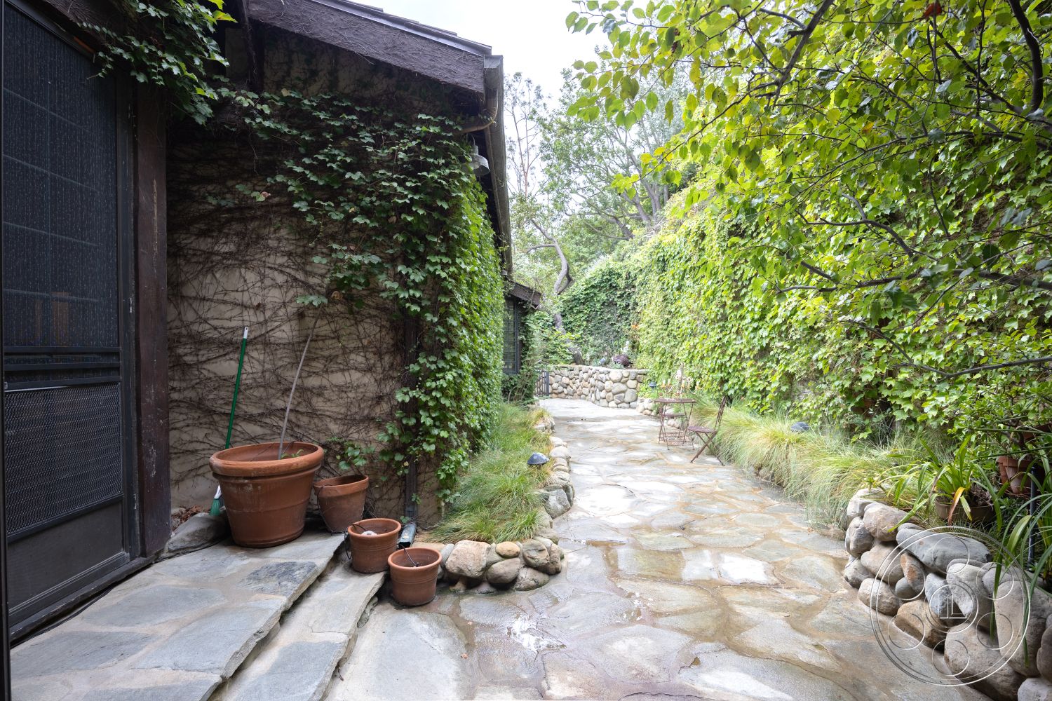 Mansion 2 - flagstone walkway, lush garden setting, exterior stone pathway, natural light exposure, rustic outdoor seating area, natural stone landscape border, exposed brick wall with ivy, wood beam roof eaves, terra cotta flower pots, greenery-clad garden wall, pebble stone garden bed, landscape grasses, private patio space, transitional outdoor area
