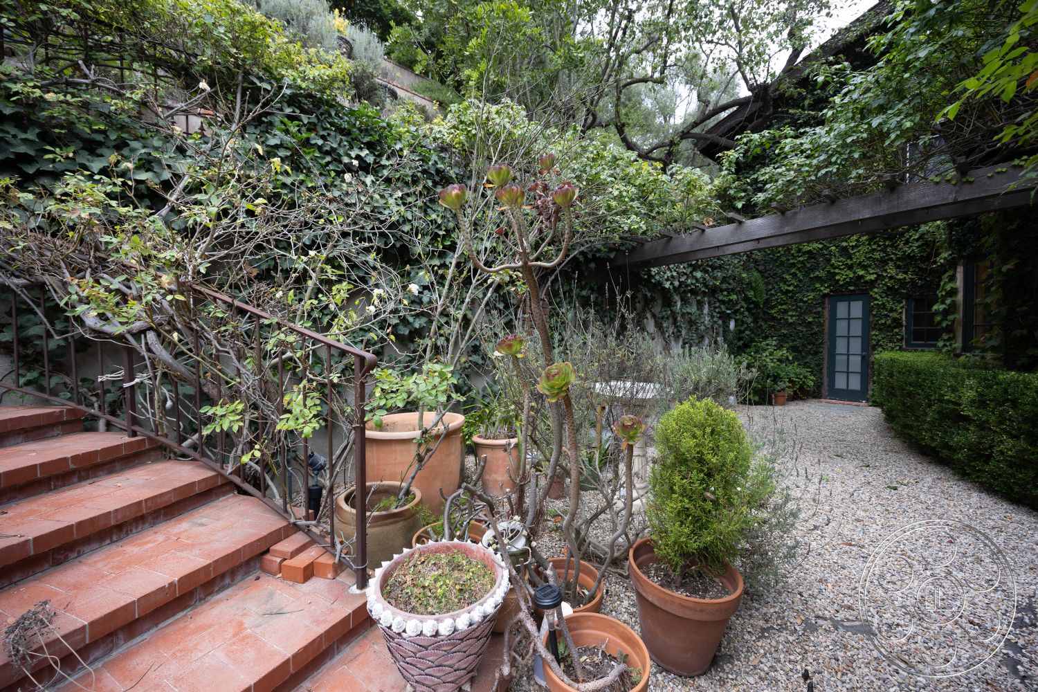 Mansion 2 - low-maintenance landscaping, lush greenery, textured stone wall, potted plants, wrought iron railing, gravel patio flooring, integrated seating area, natural light exposure, exterior garden setting, brick staircase, wooden trellis feature, earth-toned terracotta pots, rustic garden charm