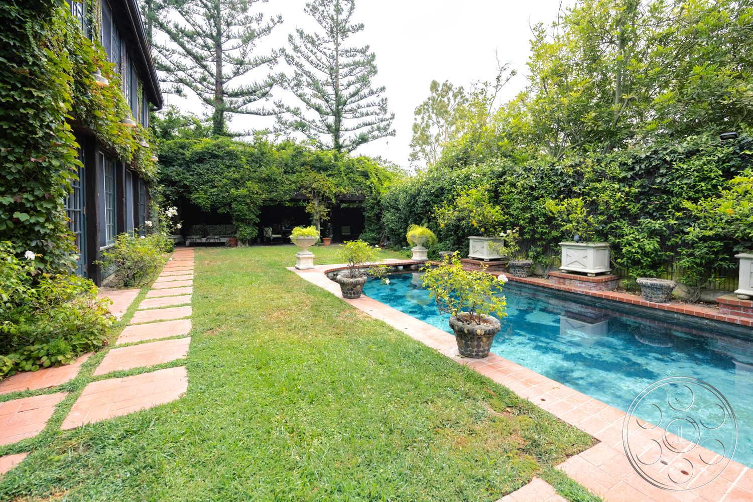 Mansion 2 - rectangular swimming pool, exposed brick wall, wooden outdoor furniture, lush green landscaping, concrete pool coping, stone planter boxes, grassy lawn area, exterior brick patio pavers, framed garden beds, tropical tree backdrop, shade provided by foliage, surrounding hedge privacy, outdoor relaxing area, garden seating zone, sun-dappled yard space