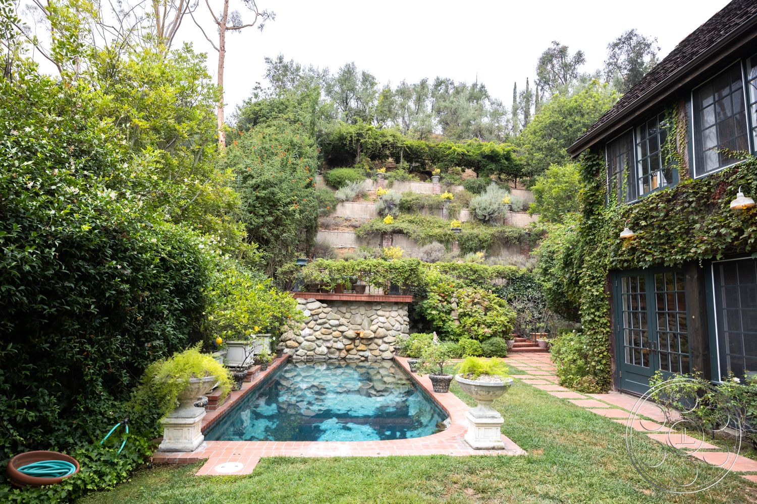 Mansion 2 - rectangular swimming pool, textured stone pathway, ivy-covered wall, stone flower bed border, lush garden landscaping, urban garden oasis, vaulted ceiling details, two-story residential home, exterior stone pool wall, red brick patio flooring, wooden deck stairs, hardwood window frames, natural hillside setting, historic-style architecture