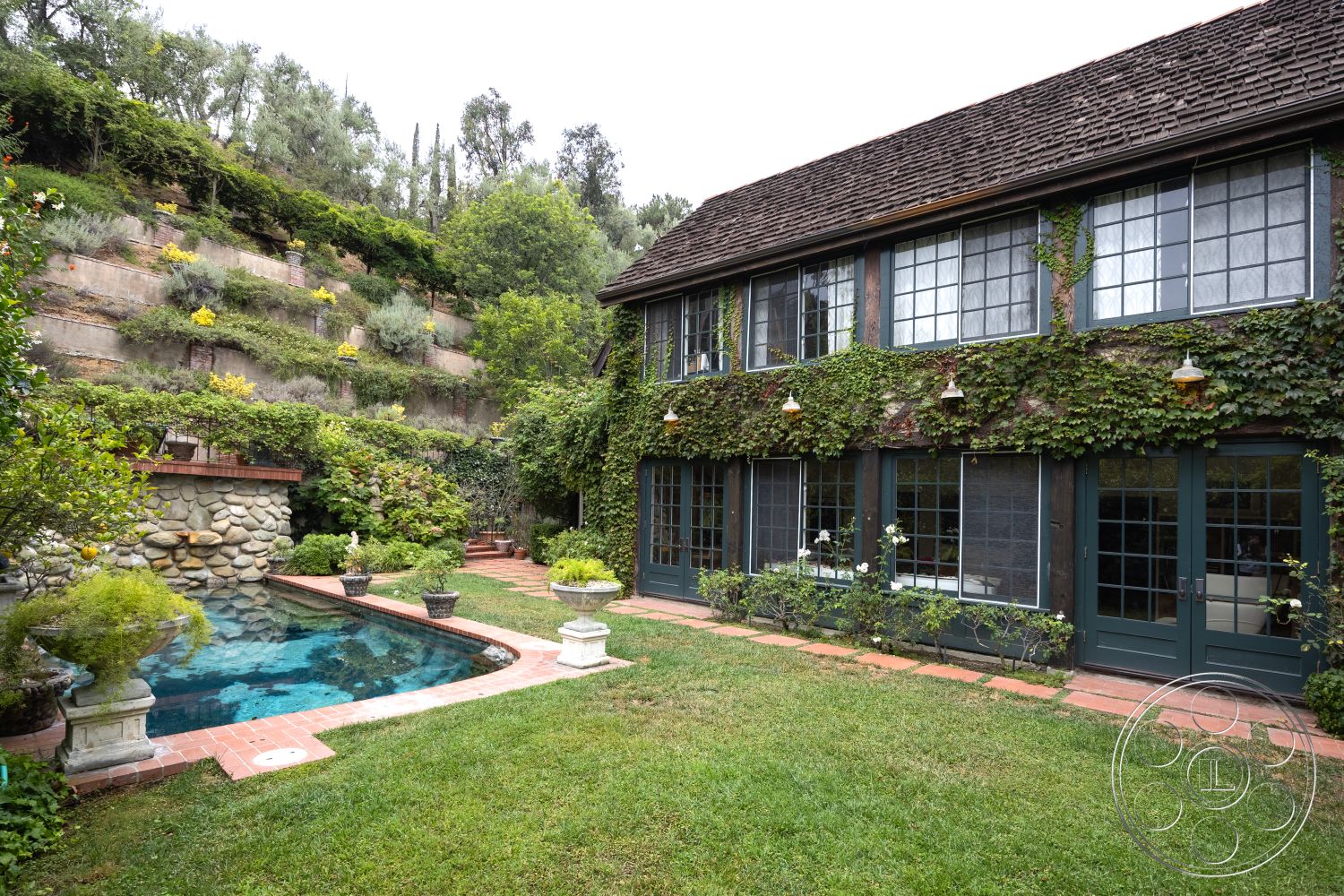 Mansion 2 - rectangular pool, double-hung windows, cobblestone walkway, wood beam ceiling, green lawn area, rustic outdoor setting, brick patio flooring, wooden deck railing, exterior stone pool border, ivy-covered exterior walls, masonry stone wall, landscape garden beds, sloped hillside setting, circular garden steps, terraced planting beds, traditional garden fountain, white frame cottage windows, classic gabled roof, patio with garden plantings