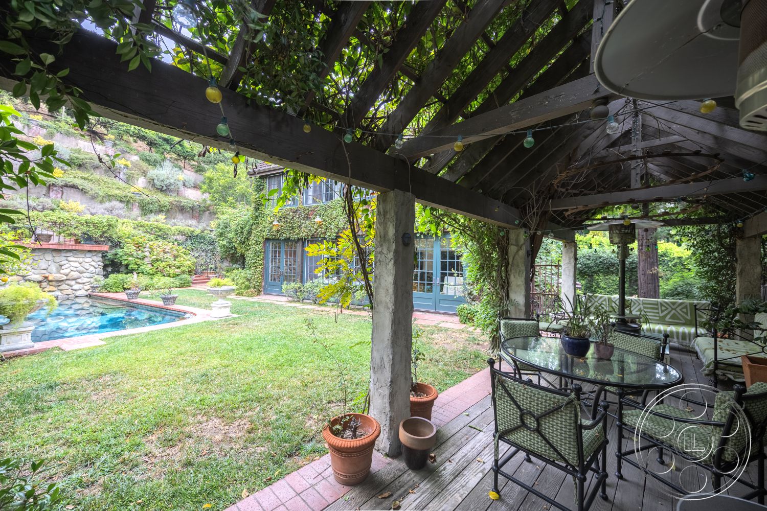 Mansion 2 - rectangular swimming pool, exposed wood beams, brick walkway, concrete planters, decorative string lights, lush green landscaping, urban garden setting, hardwood patio flooring, natural stone pool deck, surrounding hillside view, exterior patio with wooden pergola, concrete pool edge, stone garden walls, modern outdoor dining set, planter pots on patio, lawn with manicured grass, two-story home with blue windows, private backyard pool area, outdoor seating area under pergola, vining plants on structure
